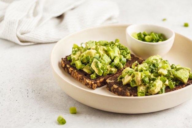 Toast à l&#39;avocat avec pain de blé entier, avocat et oignon vert sur une plaque blanche.