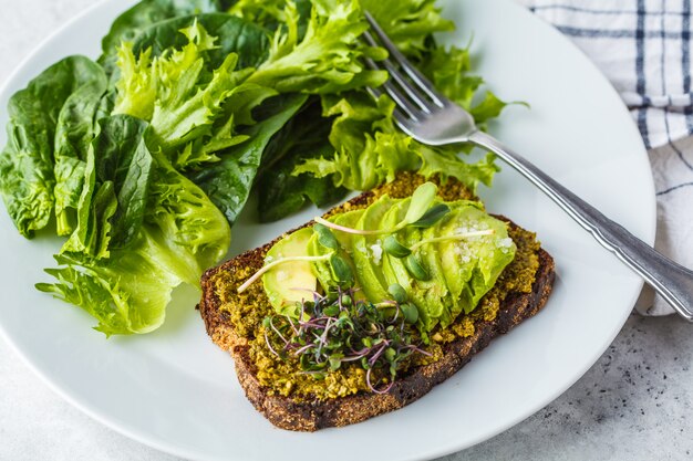 Toast à l&#39;avocat avec pesto, pousses et salade sur une plaque blanche.