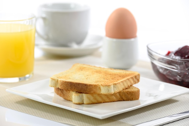 Toast croustillant sur une assiette et un œuf à la coque à côté d'une tasse de jus d'orange de café et de confiture de framboises sur fond blanc