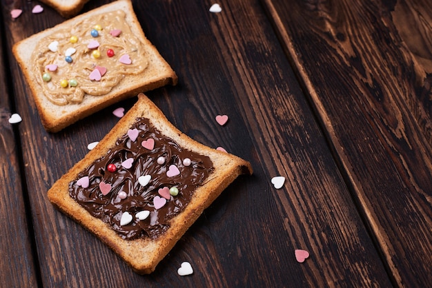 Toast avec pâte de chocolat et beurre d'arachide saupoudré de coeurs colorés sur fond de bois foncé, dessert sucré.