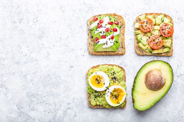 Toasts à l'avocat avec œuf, tomates, assaisonnements et moitié d'avocat entier sur fond de pierre blanche, espace pour le texte. Sandwichs sains à l'avocat pour le petit-déjeuner avec différentes garnitures, vue de dessus, gros plan