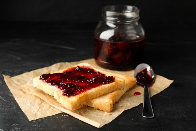 Toasts et bocal en verre avec de la confiture, une cuillère et du papier sulfurisé sur fond noir, Close up