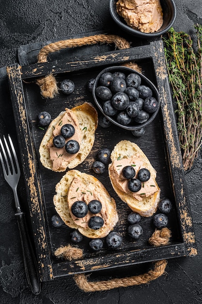 Toasts de foie gras, pâté de foie de canard et myrtille fraîche dans un plateau en bois. Fond noir. Vue de dessus.