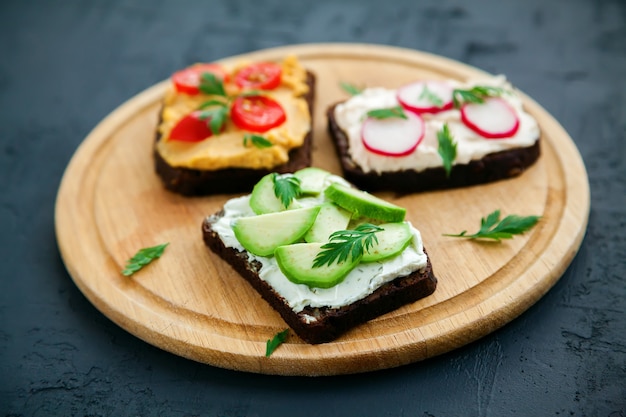 Toasts de pain de seigle végétarien avec fromage cottage, houmous, avocat, radis et tomate sur une planche de bois, surface noire