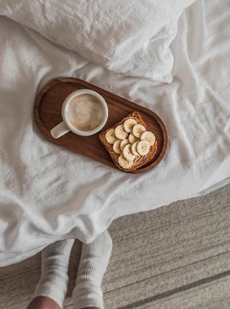 Toasts avec pâte d'arachide et cappuccino de banane sur un plateau en bois sur le lit pieds de femmes dans des chaussettes chaudes Une matinée confortable