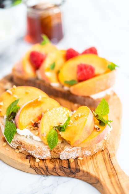 Toasts de ricotta aux pêches garnis de noix, de menthe fraîche et de framboises sur une planche à découper en bois.