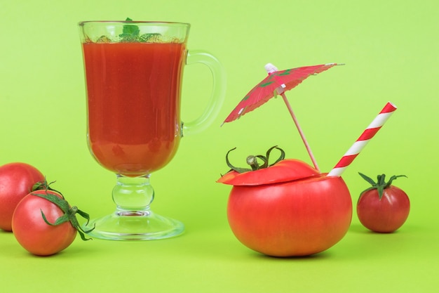 Tomate tranchée avec un tube à cocktail, un verre de jus de tomate et des tomates éparses sur fond vert.