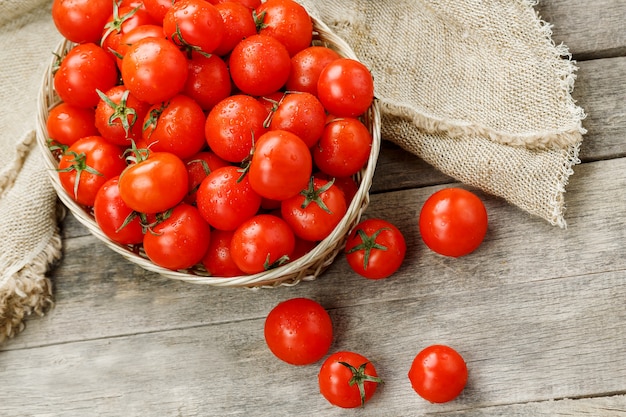 Tomates cerises mûres et juteuses avec des gouttes d'humidité dans un panier en osier. Vieille table en bois, autour de la toile de jute