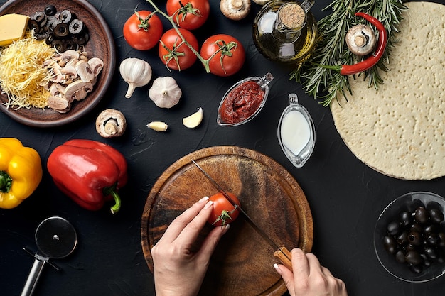 Tomates coupées à la main féminines sur une planche de bois sur la table de la cuisine autour des ingrédients du mensonge pour les légumes à pizza