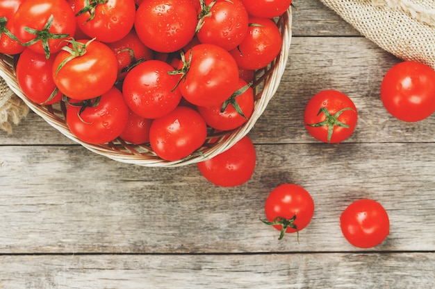 Tomates rouges fraîches dans un panier en osier sur une vieille table en bois. Tomates cerises mûres et juteuses avec des gouttes d'humidité, table en bois gris, autour d'un chiffon de toile de jute. Dans un style rustique.