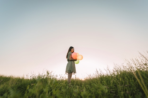 Ton film de femme asiatique solitaire tenant des ballons colorés sur un champ vert avec l'heure du coucher du soleil