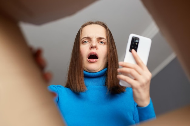 Étonné caucasienne jeune femme adulte portant un col roulé bleu ouvrant une boîte en carton regardant à l'intérieur de l'emballage avec un visage choqué tenant un téléphone intelligent à la main se tromper vue de colis du bas