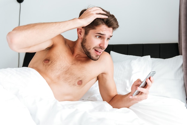 Étonné, choqué, jeune homme utilisant un téléphone portable au lit le matin