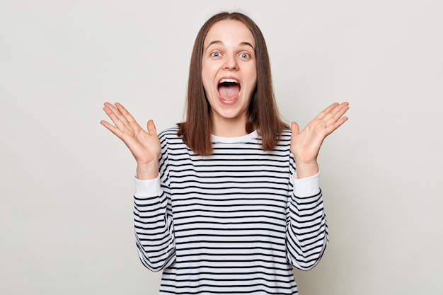 Étonné excité femme étonnée aux cheveux bruns portant une chemise rayée debout isolé sur fond gris leva les bras en criant d'émotions positives