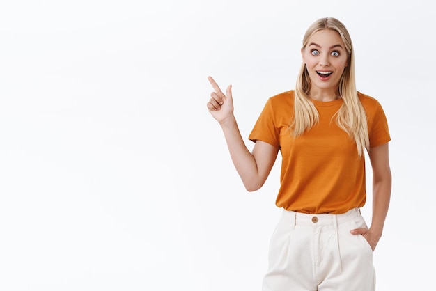 Étonné fasciné beau blond enthousiaste femme moderne en t-shirt orange haletant étonné bouche ouverte impressionné pointant le coin supérieur gauche réagir à de merveilleuses nouvelles cool