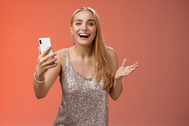 Étonné heureux charmant fille blonde en robe élégante scintillante d'argent tenant le smartphone étonné d'aimer le résultat impressionnant modifier l'application de photo souriant se demande amusé, debout fond rouge.