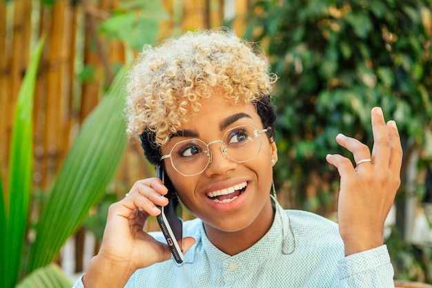 Étonné jolie fille afro-américaine aux cheveux bouclés blonds bavardant avec un ami par téléphone au café tropical