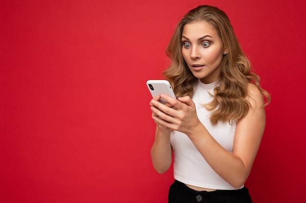 Étonnée, choquée, belle jeune femme blonde portant un t-shirt blanc isolé sur fond rouge à l'aide d'un smartphone et discutant via un téléphone portable en regardant l'écran gadjet.