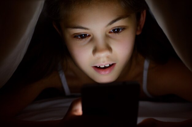 Étonnée, choquée, la fillette regarde son téléphone portable sous la couverture la nuit. Petite fille est surprise de jouer à des jeux sur son smartphone