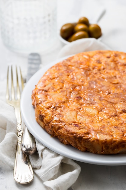 Tortilla de cuisine espagnole traditionnelle avec pommes de terre sur plat blanc et olives dans un petit bol sur fond en céramique.