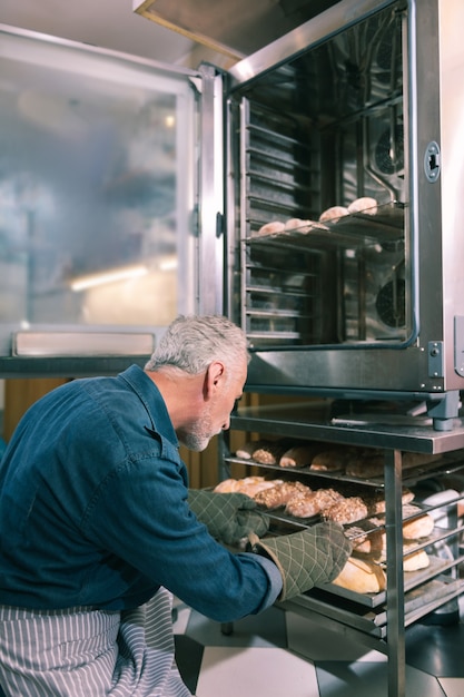 Tôt le matin. Propriétaire barbu de boulangerie mettant des croissants dans le four travaillant tôt le matin