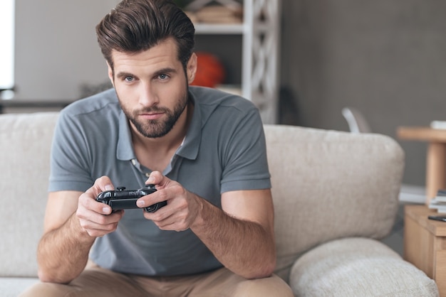 Totalement concentré sur son jeu. Beau jeune homme jouant au jeu vidéo assis sur le canapé à la maison