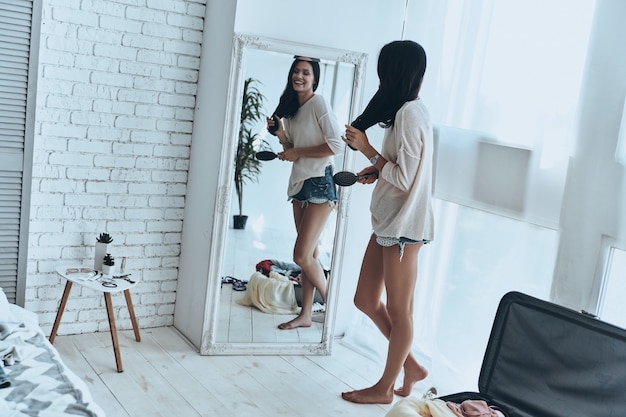 Totalement prêt. Toute la longueur d'une jeune femme séduisante combinant ses cheveux et souriant tout en se tenant près du miroir à la maison