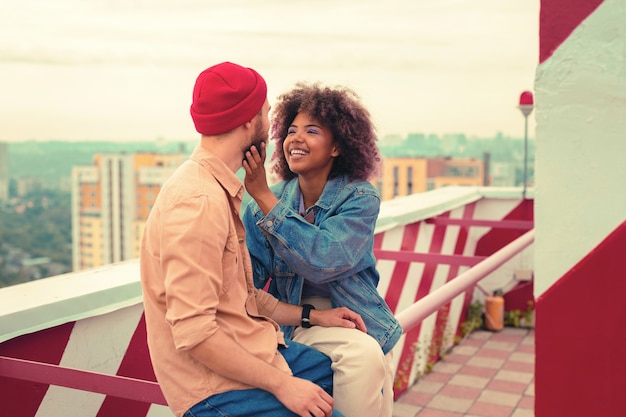Photo toucher le visage. enthousiaste jeune femme émotionnelle souriante assise sur le toit avec son petit ami et en touchant son visage