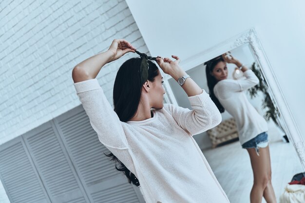 Toujours à la mode. Jolie jeune femme essayant un bandana en se tenant debout près du miroir à la maison