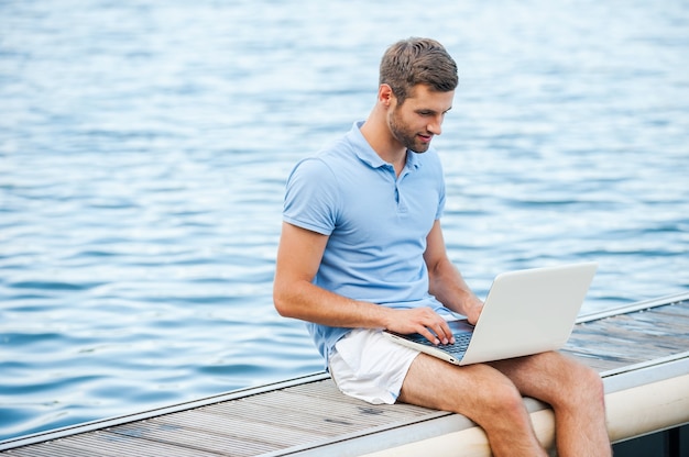 Toujours rester en contact. Beau jeune homme en polo travaillant sur ordinateur portable assis sur le quai