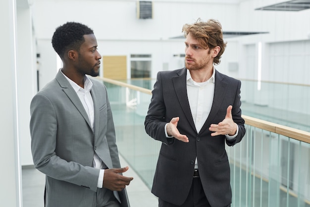 Tour De Taille Portrait De Deux Hommes D'affaires Prospères Portant Des Costumes En Discutant Tout En Marchant Vers La Caméra Dans L'intérieur D'un Immeuble De Bureaux Moderne