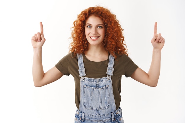 Tour de taille sympathique rousse ambitieuse bouclée montre la promo supérieure, pointant vers le haut, souriante ravie, présente la publicité, mur blanc debout dans une salopette en jean et un t-shirt