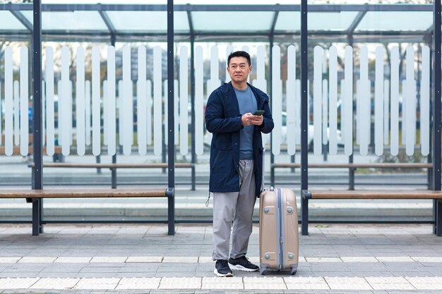 Touriste d'affaires asiatique, attendant un taxi près de l'aéroport, utilise l'application téléphonique pour louer une voiture, un homme avec une grosse valise