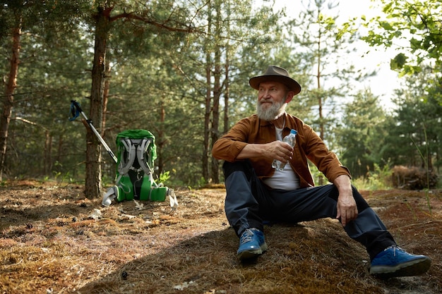 Touriste âgé pensif buvant de l'eau d'une bouteille en plastique assis sur l'herbe sèche de la forêt Voyage de survie et randonnée dans la forêt