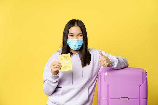 Une touriste asiatique souriante en masque facial, debout avec une valise, montrant un certificat de vaccination international covid pour les voyageurs et le pouce levé, fond jaune.