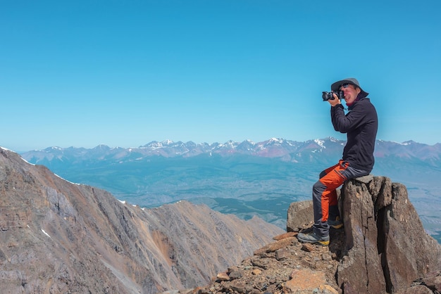 Touriste assis sur une pierre près du bord de l'abîme à haute altitude sous un ciel bleu par temps ensoleillé Homme avec caméra sur un rocher élevé près du bord du précipice avec une vue magnifique d'en haut sur une grande chaîne de montagnes