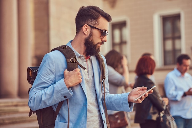 Un touriste avec une barbe complète et une coupe de cheveux, portant des vêtements décontractés et des lunettes de soleil, tient un sac à dos et envoie des SMS sur un smartphone, debout dans une rue antique, pendant l'excursion en Europe.