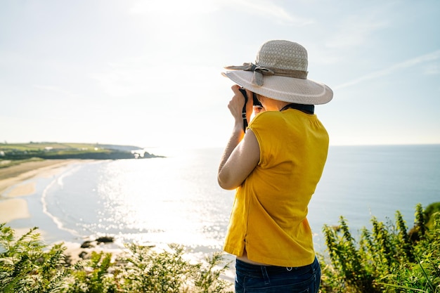 Touriste blonde capturant une vue côtière vibrante avec la lumière de midi dans un t-shirt jaune décontracté et un chapeau coloré