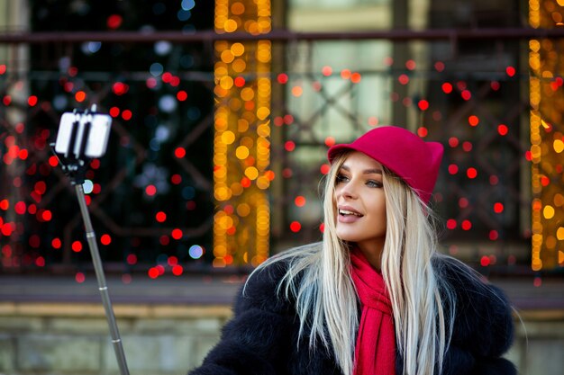 Touriste blonde glamour portant un chapeau drôle, prenant un selfie dans une rue de la ville décorée de guirlandes. Espace pour le texte