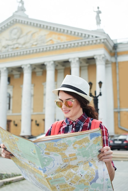 La touriste avec chapeau et lunettes de soleil tenant une carte et la regardant contre le bâtiment avec des colonnes