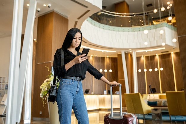 Touriste féminin sérieux avec la valise regardant par le message de téléphone