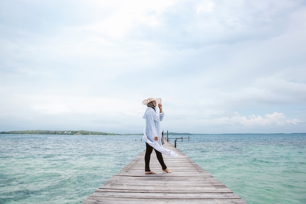 Touriste de femme dans la robe blanche et le chapeau d'été posant sur le dock en bois avec le fond de paysage marin