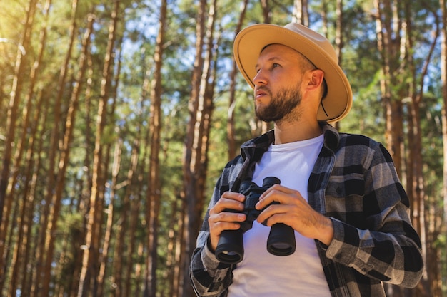 Touriste homme dans un chapeau et une chemise à carreaux regarde à travers des jumelles dans la forêt.