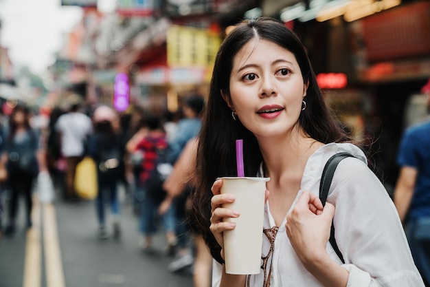 Une touriste japonaise asiatique amusante et mignonne ayant une boisson boba froide dans une tasse en papier tout en se tenant dans la rue du marché en plein air par une journée ensoleillée. belle voyageuse savourez un délicieux thé au lait à bulles avec de la paille