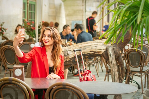 Touriste jeune femme caucasienne dans une veste rouge avec valise prend un selfie à la table du café