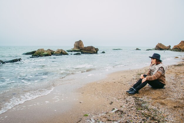 Touriste de jeune femme dans le chapeau se reposant sur la plage regardant la mer sur le littoral à l'horizon