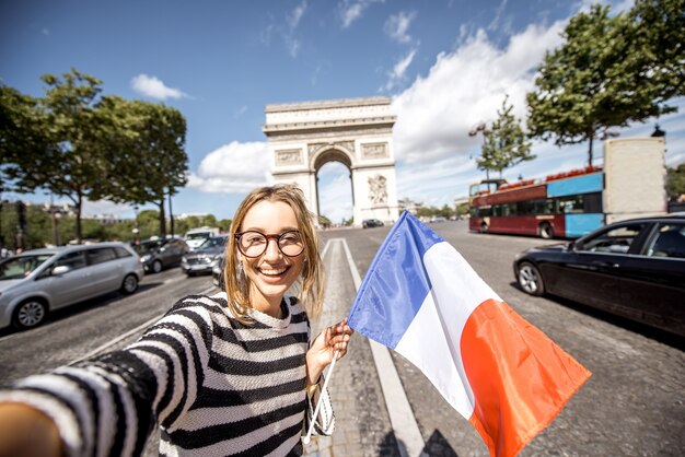 Photo touriste de jeune femme avec le drapeau français faisant le portrait de selfie avec l'arc de triomphe célèbre sur l'arrière-plan à paris