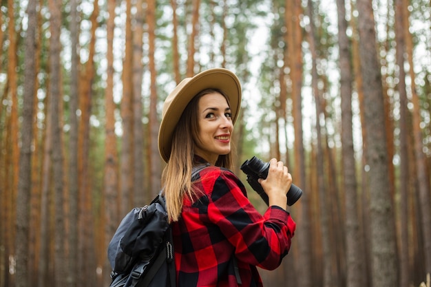 Touriste de jeune femme avec sac à dos, chapeau et chemise à carreaux rouge tenant des jumelles dans la forêt.