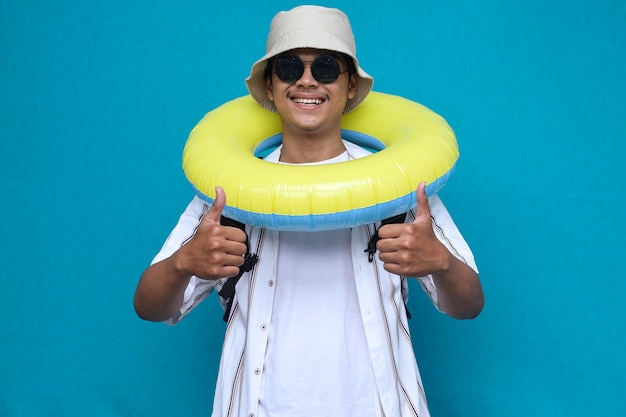 Un touriste joyeux souriant porte un chapeau de chemise de plage et tient un anneau gonflable tout en donnant le pouce vers le haut est