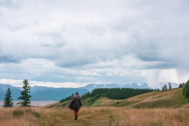 Touriste marche à travers les collines et la forêt vers le mauvais temps Randonneur sur le chemin de la grande chaîne de montagnes enneigée sous un ciel nuageux pluvieux Homme en imperméable dans les montagnes dans le nuageux Voyageur va vers l'aventure